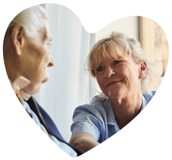 A nurse in a light blue uniform and an older woman in a dark blue top smile at each other
