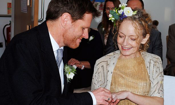 Tom placing a ring on Penny's finger during the marriage ceremony. He is wearing a suit, Penny is wearing a gold dress and cream, embroidered jacket.
