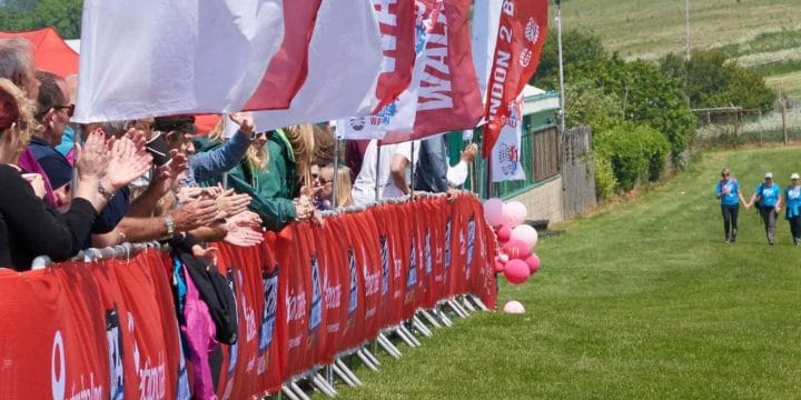 a crowd of spectators watching a group of walkers holding hands, walking along a grassy field
