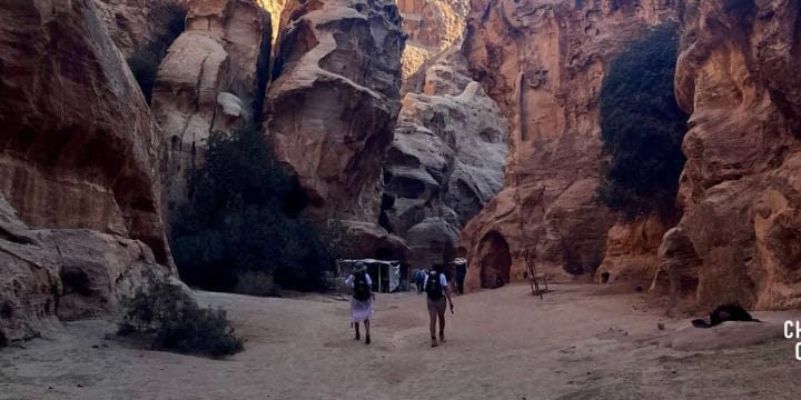 two people in hiking gear walking towards large rock formations under a bright blue sky