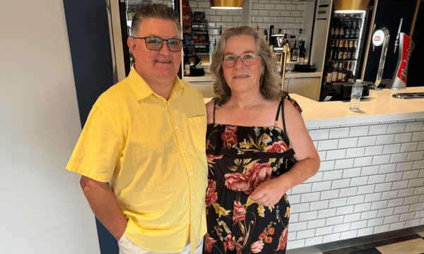 Graham and Janet standing together, wearing holiday attire. Graham is wearing a yellow shirt, Jan has a flowery dress on.