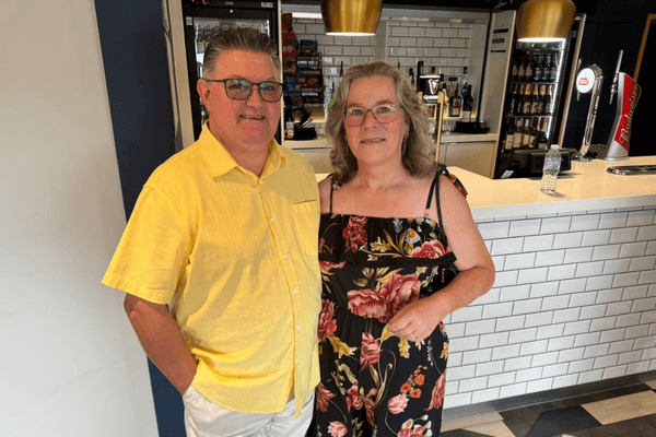 Graham and Janet standing together, wearing holiday attire. Graham is wearing a yellow shirt, Jan has a flowery dress on.