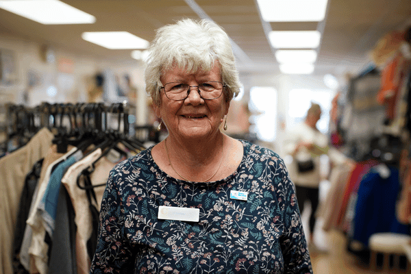 Lis is in her seventies, has white short hair and is wearing a blue, white and black flowery top. She's standing in front of clothes rails in hte shop.