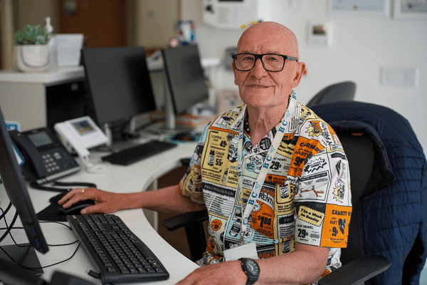 John has a bald head and glasses. he's wearing a colourful orange, yellow and white shirt and sitting at a reception desk.