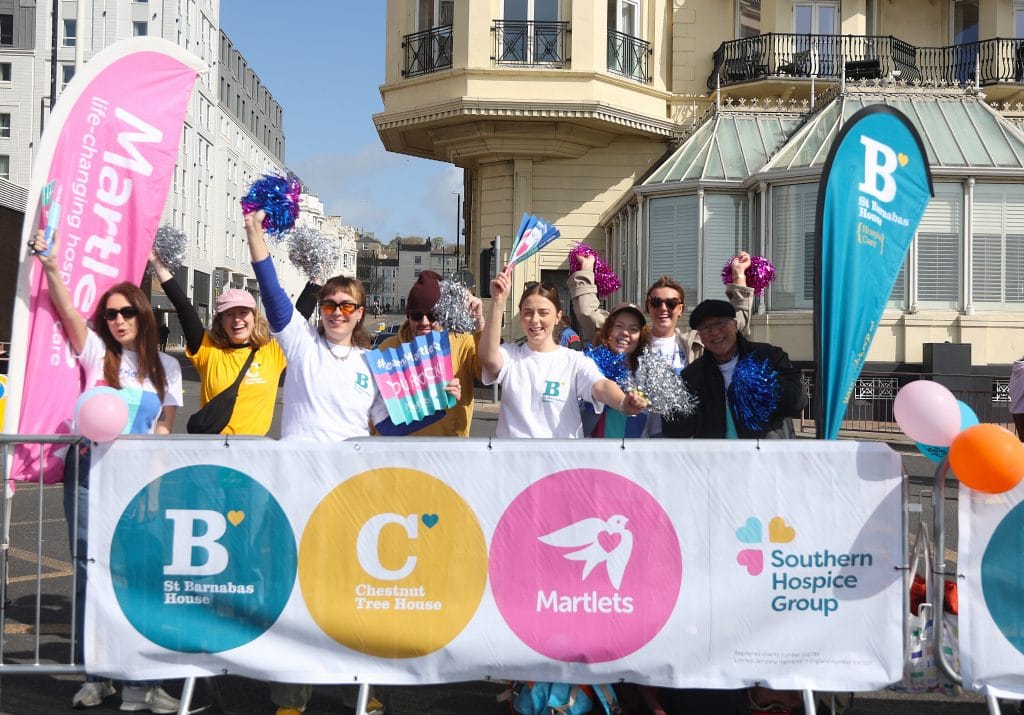 A group of supporters stands at the side of the road smiling and cheering for marathon runners