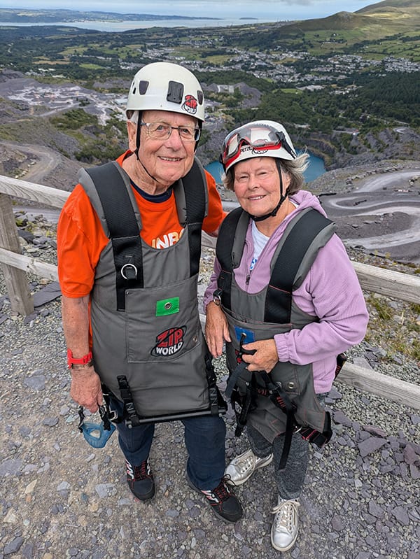 Colin and Angela in safety helmets and harnesses, standing high up overlookking a countryside view. They both smile at the camera