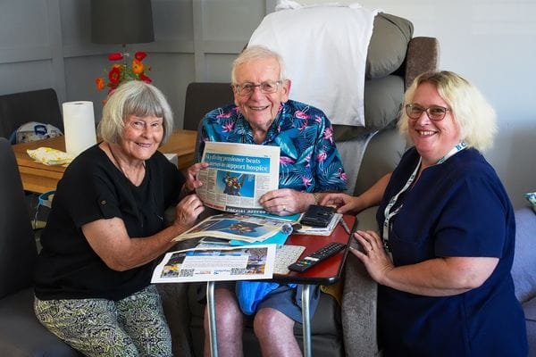 Colin, angela and julie sitting together smiling at the camera. Colin is holding up a newspaper cutting showing an image of skydiving and headline "Skydiving pensioner beats his nerves to support hospice"