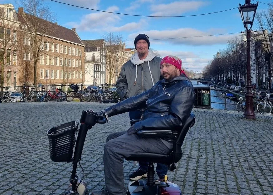 Dan and Carl on holiday by a canal in Amsterdam, Dan is in a mobility scooter